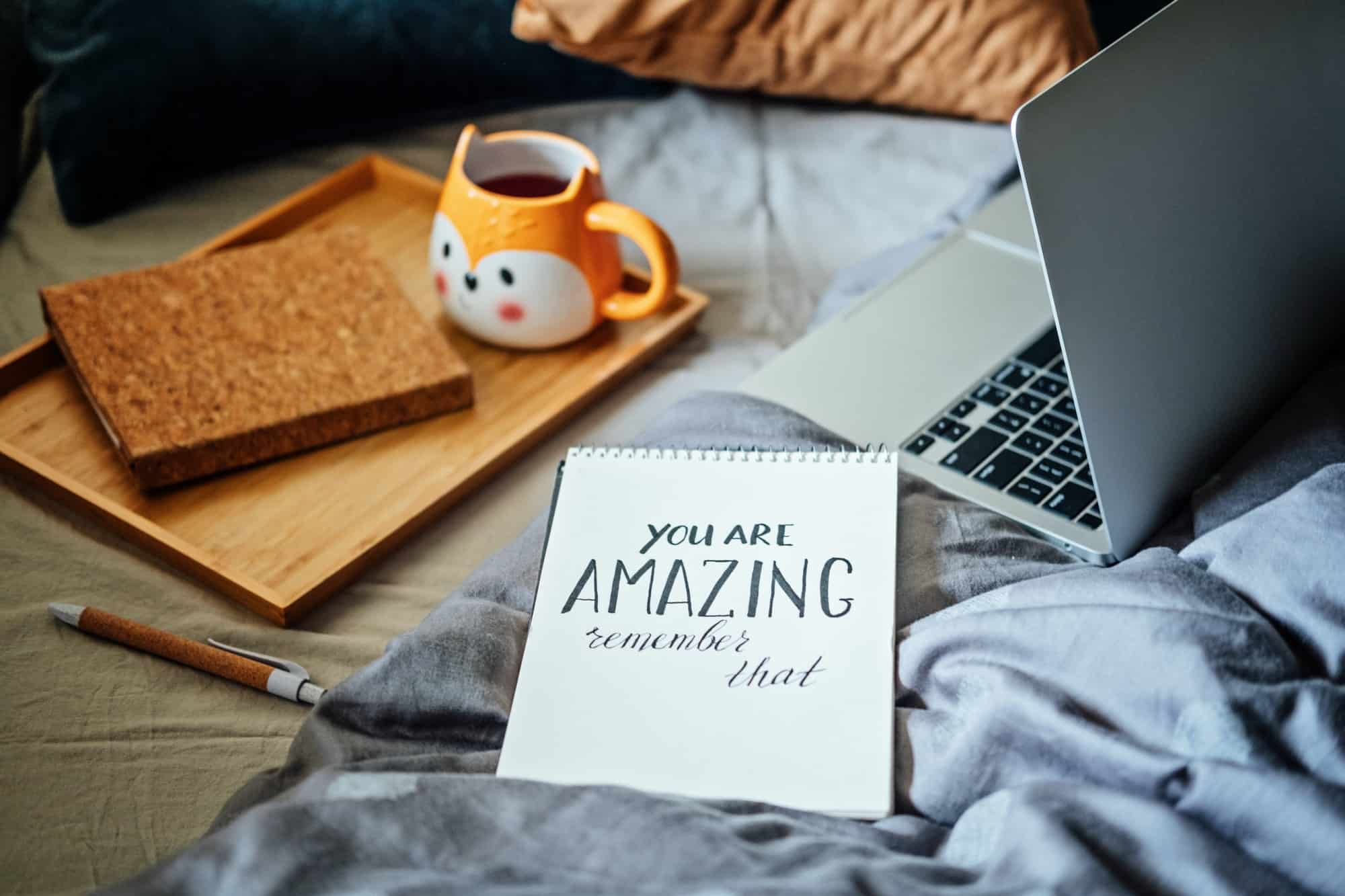 Positive daily affirmations for self love. Words You Are Amazing in notebook near laptop and tea